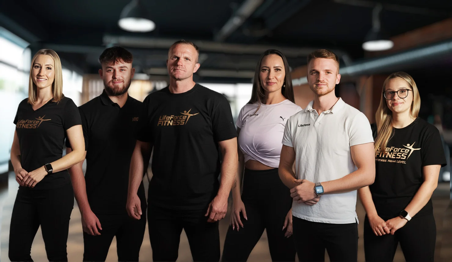 Gruppenfoto des Fitnessstudio-Teams: Sechs Personen stehen nebeneinander in einem modernen Trainingsraum und tragen LifeForce-Fitness-Shirts bzw. Sportbekleidung.