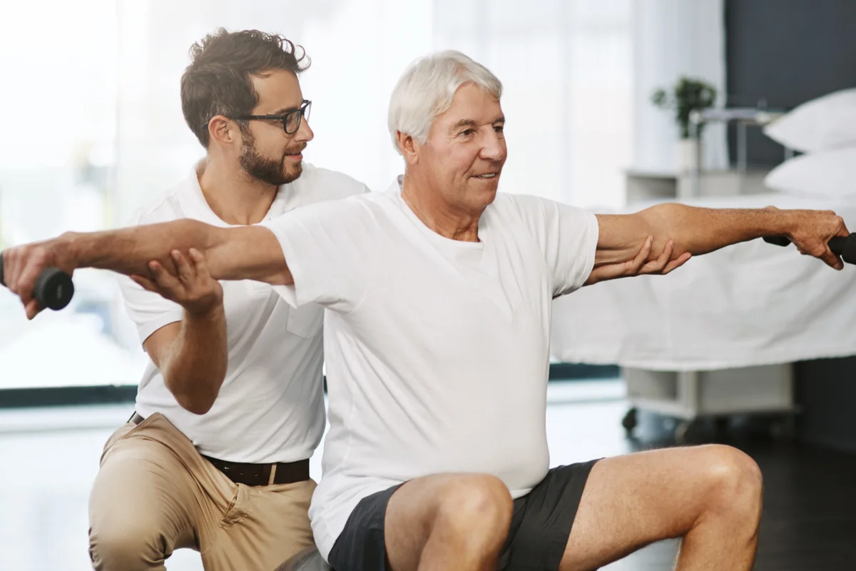 Ein älterer Mann trainiert mit ausgestreckten Armen auf einem Gymnastikball, während ein Physiotherapeut ihn seitlich unterstützt und anleitet.
